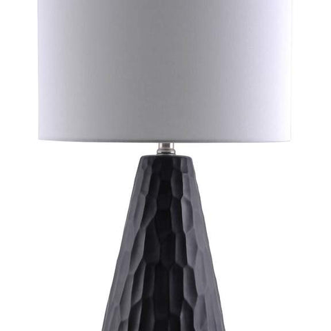 An isolated lamp with a dark base. Indented geometric patterns in the lamp base adds subtly and texture to any space. A plain white lampshade offers a point of contrast.